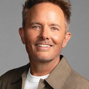 Smiling man with short brown hair wearing a beige jacket over a white shirt, posed against a gray background.